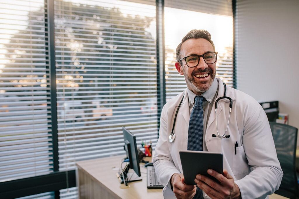 A medical professional leaning against his desk smiles while holding a tablet - Stroudwater